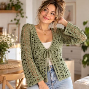 May include: A sage green crochet cardigan featuring long, flared sleeves, a deep V-neckline, and a front tie closure. The garment is crafted with an openwork, geometric stitch pattern and is worn over a white top and light blue denim jeans.