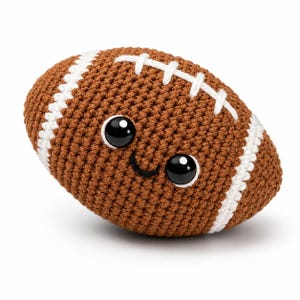 May include: A brown crochet football with white stitched laces and stripes. The football features two large, glossy black safety eyes and a small, embroidered black smile, giving the plush toy a friendly, character-like appearance.