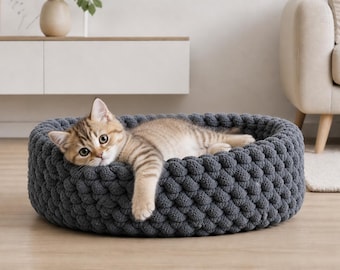 May include: A grey, round cat bed with a textured, woven design. A light brown tabby cat is lying inside the bed, looking at the camera. The bed is on a wooden floor.