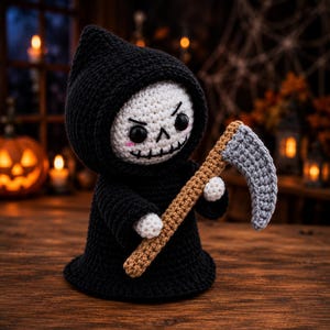 May include: A crocheted grim reaper doll featuring a black hooded cloak, a white skull face with black stitched features, pink cheek accents, and small white hands holding a scythe with a brown handle and a textured grey blade.