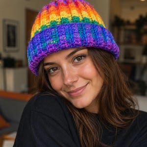 Può includere: Un berretto all'uncinetto con una trama a coste e un vivace gradiente di colori arcobaleno. Il cappello sfuma dal rosso, arancione e giallo sulla corona al verde, blu e viola sul bordo ripiegato, realizzato con un filato spesso e corposo.