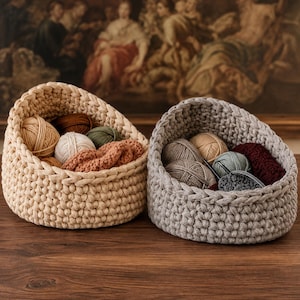 May include: Two round, hand-crocheted baskets made from thick, chunky yarn. One basket is a light beige colour, and the other is a soft grey. Both baskets are filled with various balls of yarn in neutral and earthy tones, including tan, white, olive, and dark red.