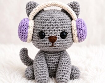 May include: A grey crocheted cat doll featuring large black eyes and a brown nose. The cat is wearing a pair of headphones with a cream-coloured headband and light purple ear cups. The entire figure is crafted with a visible, textured yarn stitch.