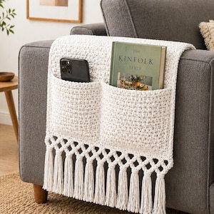 Op de afbeelding: Een crèmekleurige, gehaakte armleuningorganizer met twee zakken en een franjebies. Een zwarte smartphone en een groen boek met de titel "The Kinfolk Table" zitten in de zakken. De organizer is over de arm van een grijze bank gedrapeerd.