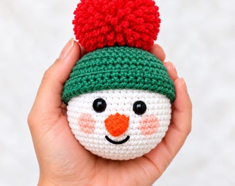 May include: A round, white crocheted snowman head ornament featuring a green beanie with a large red yarn pom-pom on top. The snowman has two black plastic eyes, a small orange crocheted carrot nose, and a stitched black smile with pink cheeks.