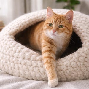 May include: An orange tabby cat emerges from a cream-colored, knitted cat bed. The cat's front paw is extended, and its green eyes are visible. The cat bed is round and has a cozy, enclosed design, perfect for a pet.