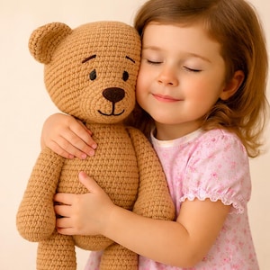 Op de afbeelding: Een gehaakte teddybeer in bruintinten wordt omhelsd. De beer heeft een donkerbruine neus en zwarte ogen. De beer wordt vastgehouden door een persoon die een roze bloemenjurk draagt.