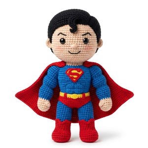 Könnte beinhalten: Eine handgefertigte Häkelpuppe, die einen Superhelden mit schwarzem Haar, blauem Anzug, roten Stiefeln und einem roten Umhang darstellt. Die Figur hat einen gelben Gürtel, ein rot-gelbes Brustemblem und einen hellen Hautton mit rosa Wangen.