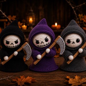 May include: Three crocheted figures dressed in hooded robes of black, purple, and dark gray. Each figure has a white face with black eyes, a stitched mouth, and pink cheeks. They each hold a small, light brown crocheted scythe with a gray blade.