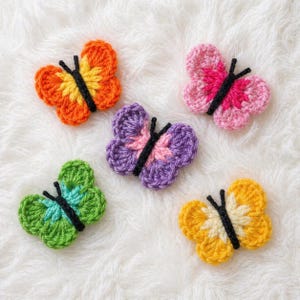 May include: Five colorful crochet butterfly appliques. The butterflies feature various color combinations, including orange and yellow, pink and magenta, purple and pink, green and blue, and yellow and cream. Each butterfly has black antennae.
