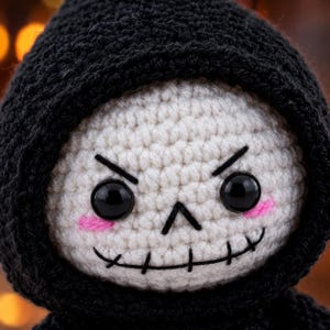 May include: A crocheted doll featuring a white skull-like face with black stitched eyebrows, a stitched mouth, and round black plastic eyes. The doll wears a textured black crocheted hood that frames the face, with small pink stitched cheeks.