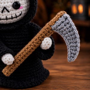 May include: A crocheted figure of a grim reaper wearing a black hooded robe with white hands and a white skull face featuring black eyes, a stitched mouth, and pink cheeks. The figure holds a crocheted scythe with a brown handle and a gray blade.