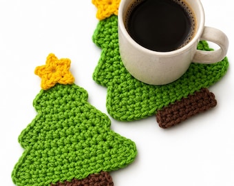 May include: Two handmade crochet coasters shaped like Christmas trees. Each coaster features a green triangular tree body, a brown rectangular base, and a bright yellow star at the top. One coaster supports a ceramic mug filled with black coffee.