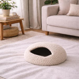 May include: A cream-colored, oval pet bed with a dark interior. The bed is made of a thick, textured material and sits on a light-colored rug. The pet bed is designed to provide a cozy space for a small animal.