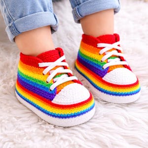 May include: A pair of handmade crochet baby shoes in a rainbow color scheme. The shoes have a red upper, white soles, and white laces. The colorful design includes stripes of red, orange, yellow, green, blue, and purple. The shoes are on a white fluffy surface.