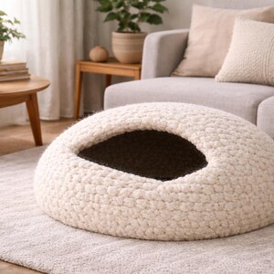 May include: A cream-colored, dome-shaped pet bed with a large opening. The bed is made of a thick, textured material and sits on a light gray rug. The interior of the bed is dark. The scene includes a sofa, coffee table, and plants.