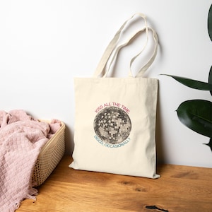Pode incluir: Bolsa tote creme com um gráfico de bola de discoteca e o texto "KISS ALL THE TIME" em rosa e "DISCO, OCCASIONALLY" em verde-azulado. A bolsa tem alças longas e é feita de tecido resistente.