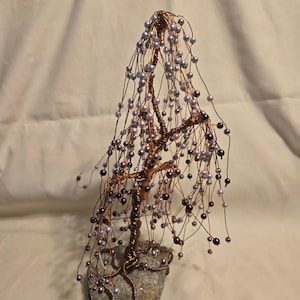 May include: A handcrafted wire tree sculpture with copper-colored branches and cascading strands of beads in shades of gray, purple, and black. The tree is mounted on a gray stone base, creating a unique decorative piece.