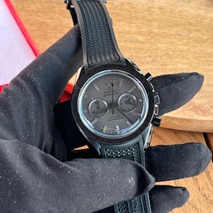 Puede incluir: Un reloj Omega Speedmaster negro con esfera gris y correa de caucho negro. El reloj tiene subesferas de cronógrafo y una pantalla de fecha. El reloj está sujeto por una mano enguantada.