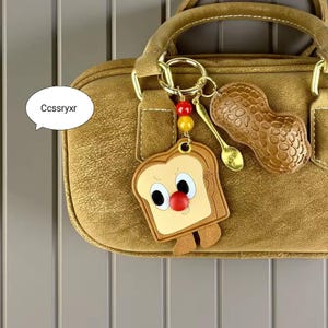 Puede incluir: Un bolso marrón con detalles dorados, con un llavero con un dije de rebanada de pan de dibujos animados, maní y cuchara. La rebanada de pan tiene una cara con una nariz roja. El texto "Ccssryxr" está en un bocadillo.