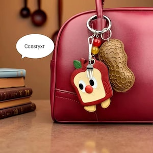 Puede incluir: Un bolso de mano de cuero rojo con un llavero plateado que sostiene un llavero de dibujos animados de manzana y cacahuete. El llavero de manzana tiene un diseño de tenedor y el texto "BWSPN". El texto "Ccssryxr" está en una burbuja de diálogo blanca.