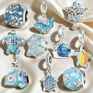 May include: A collection of silver charm beads with ocean-themed designs. The charms include turtles, a whale, a starfish, a snail, and a seashell, all in shades of blue, white, and silver. The beads are displayed on a satin fabric.