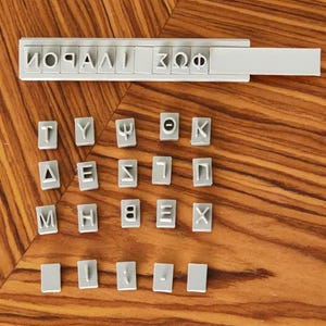 May include: A collection of grey letter stamps and a stamp holder, featuring Greek letters. The individual letter stamps are arranged on a wooden surface. The stamp holder has Greek letters printed on it.