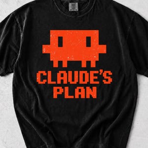 May include: A black short-sleeve t-shirt featuring a distressed orange pixelated graphic of a square-shaped character above the text CLAUDE'S PLAN in a matching orange pixelated font. The shirt has a crew neckline and a standard fit.