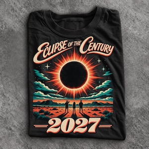May include: A black t-shirt featuring a graphic of a solar eclipse above a desert landscape with three figures standing in the center. The text reads "ECLIPSE OF THE CENTURY" at the top and "2027" at the bottom in a bold, retro-style font.