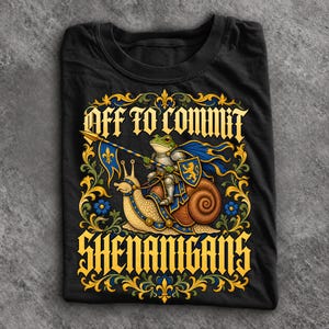 May include: A black t-shirt featuring a graphic of a frog knight in silver armour riding a large snail. The frog holds a blue and gold spear and shield. The text reads OFF TO COMMIT SHENANIGANS in a yellow gothic font, framed by ornate gold and blue floral scrollwork.