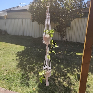 May include: A white macrame plant hanger with two beige pots, each holding a green leafy plant. The hanger is suspended from a metal ring, with the plants hanging in the air. The background includes a grassy lawn and a fence.