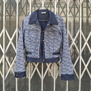 Può includere: Una giacca in denim con un motivo ripetuto della lettera m minuscola in bianco su sfondo azzurro. La giacca presenta colletto, polsini e abbottonatura frontale blu scuro con quattro bottoni metallici, due tasche sul petto e maniche lunghe.