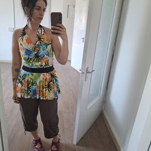 May include: A halter top featuring a vibrant floral print in shades of blue, orange, and green with a black waistband, paired with dark brown cropped trousers and dark red flat shoes. The top has a cowl neckline and a flared hemline.