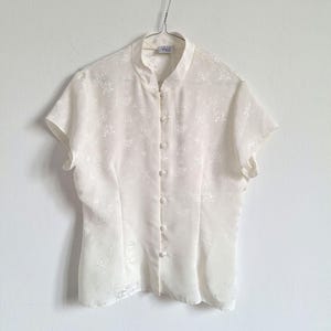 Puede incluir: Una blusa de color blanco roto con mangas cortas, cuello mao y cierre frontal de botones con cinco botones a juego cubiertos de tela. La prenda está confeccionada en un tejido semitransparente con un sutil estampado floral tonal.