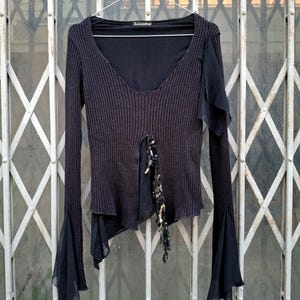 May include: Black long-sleeve top with a V-neck and flared sleeves. The top features a ribbed knit bodice with a sheer fabric overlay and embellishments down the front. The sleeves have a flared, bell-like shape. The top is hanging on a metal hanger.