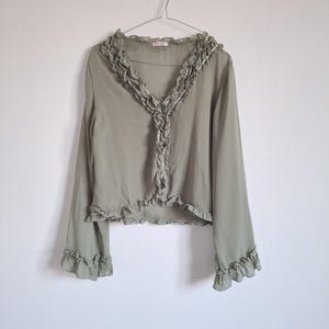 Pode incluir: Uma blusa de manga comprida em verde sálvia com decote em V e acabamento em folhos que desce pela abertura frontal. A peça apresenta mangas largas com punhos com folhos e uma bainha com folhos, feita de tecido leve e semitransparente.