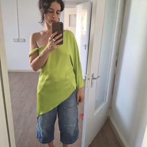 May include: A lime green one-shoulder top with a loose, asymmetrical hemline paired with blue denim cargo-style cropped pants and black platform sandals. The top features a single long sleeve and a smooth, slightly textured fabric finish.