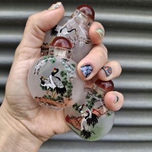Può includere: Tre piccole boccette da fiuto rotonde in vetro traslucido con tappi rosso scuro. Ogni boccetta presenta una scena dipinta a mano di gru bianche e nere con corone rosse, pini verdi e formazioni rocciose marroni, accompagnate da calligrafia nera.