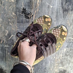 Può includere: Un paio di sneakers marroni con tessuto fantasia nei toni del verde, giallo e marrone. Le sneakers hanno lacci e cinturini marroni. Le scarpe sono tenute da una mano.