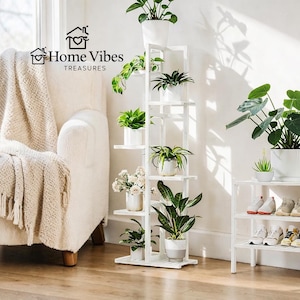 May include: A white wooden plant stand with multiple shelves, displaying various potted plants. The stand is positioned near a window and a cream-colored armchair. The Home Vibes Treasures logo is visible.