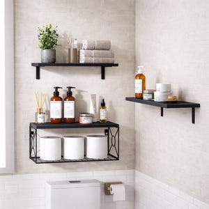 May include: A bathroom scene featuring three black shelves mounted on a light-colored brick wall. The shelves hold various toiletries, towels, and a small potted plant. The bottom shelf has a decorative metal frame and holds toilet paper rolls.