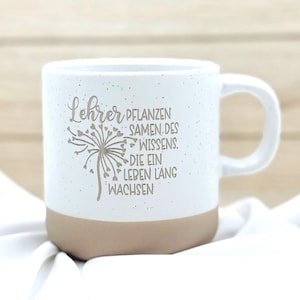 Könnte beinhalten: Ein weißer Keramikbecher mit Sprenkeloptik und beigefarbenem Boden. Auf der Vorderseite befindet sich eine braune Grafik einer Pusteblume mit herzförmigen Samen und der Text: Lehrer pflanzen Samen des Wissens, die ein Leben lang wachsen.