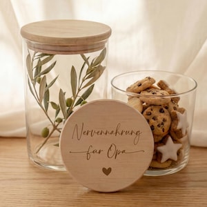 Puede incluir: Un tarro de cristal transparente con tapa de madera y ramas de olivo en su interior, junto a un vaso pequeño lleno de galletas. Una tapa de madera con el texto "Nervennahrung für Opa" y un corazón está delante.