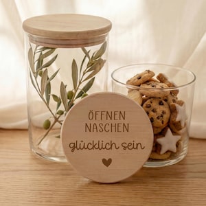 Könnte beinhalten: Ein klares Glas mit Holzdeckel, gefüllt mit Olivenzweigen, steht neben einem kleineren Glas mit Keksen. Eine Holzscheibe mit deutschem Text und einem Herz befindet sich davor.