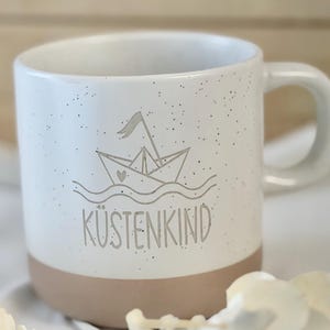 Könnte beinhalten: Weißer Keramikbecher mit gesprenkelter Oberfläche und beigefarbenem Boden. Der Becher zeigt eine graue Illustration eines Papierbootes auf Wellen, darunter das Wort "KÜSTENKIND". Der Becher hat einen gebogenen Henkel.