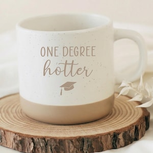 Op de afbeelding: Een gespikkelde witte keramische mok met een beige basis, voorzien van de bruine tekst ONE DEGREE hotter en een klein icoontje van een afstudeerhoed. De mok heeft een gebogen handvat en een gladde afwerking voor een strak ontwerp.