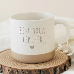 Puede incluir: Una taza de cerámica blanca con el texto "BEST YOGA TEACHER" y un corazón. La mitad inferior de la taza es de color beige. La taza está sobre un posavasos de madera.