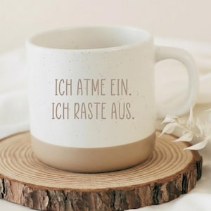 Könnte beinhalten: Eine weiße Keramik-Tasse mit gesprenkelter Oberfläche und beigefarbenem Boden, auf einem Holzuntersetzer. Die Tasse trägt den Text "ICH ATME EIN. ICH RASTE AUS." in braunen Buchstaben. Die Tasse hat einen gebogenen Henkel.