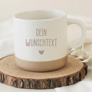 Op de afbeelding: Een witte keramische mok met een gespikkelde afwerking en een bruine basis. De mok heeft de tekst "DEIN WUNSCHTEXT" en een hartsymbool. Hij staat op een houten onderzetter, met een witte achtergrond.