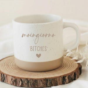 Könnte beinhalten: Eine weiße Keramik-Tasse mit gesprenkelter Oberfläche und beigefarbenem Boden. Die Tasse trägt den Schriftzug "moingiorno BITCHES" in Schreibschrift sowie ein Herzsymbol. Die Tasse steht auf einem runden Holzuntersetzer.